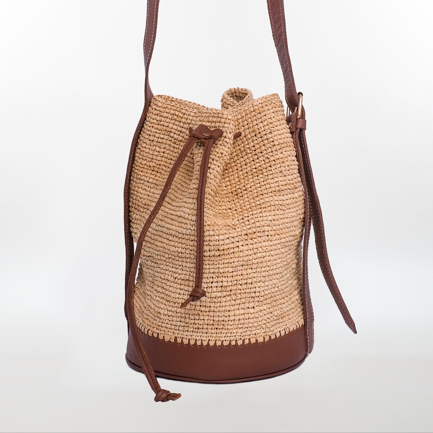 Natural raffia bucket bag with brown leather base, drawstring closure, and leather strap