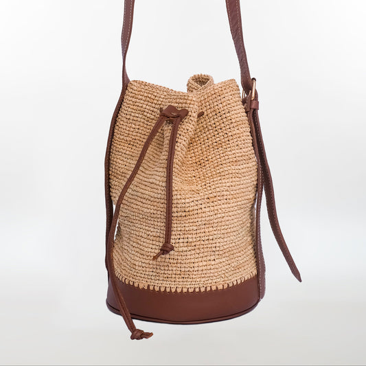 Natural raffia bucket bag with brown leather base, drawstring closure, and leather strap