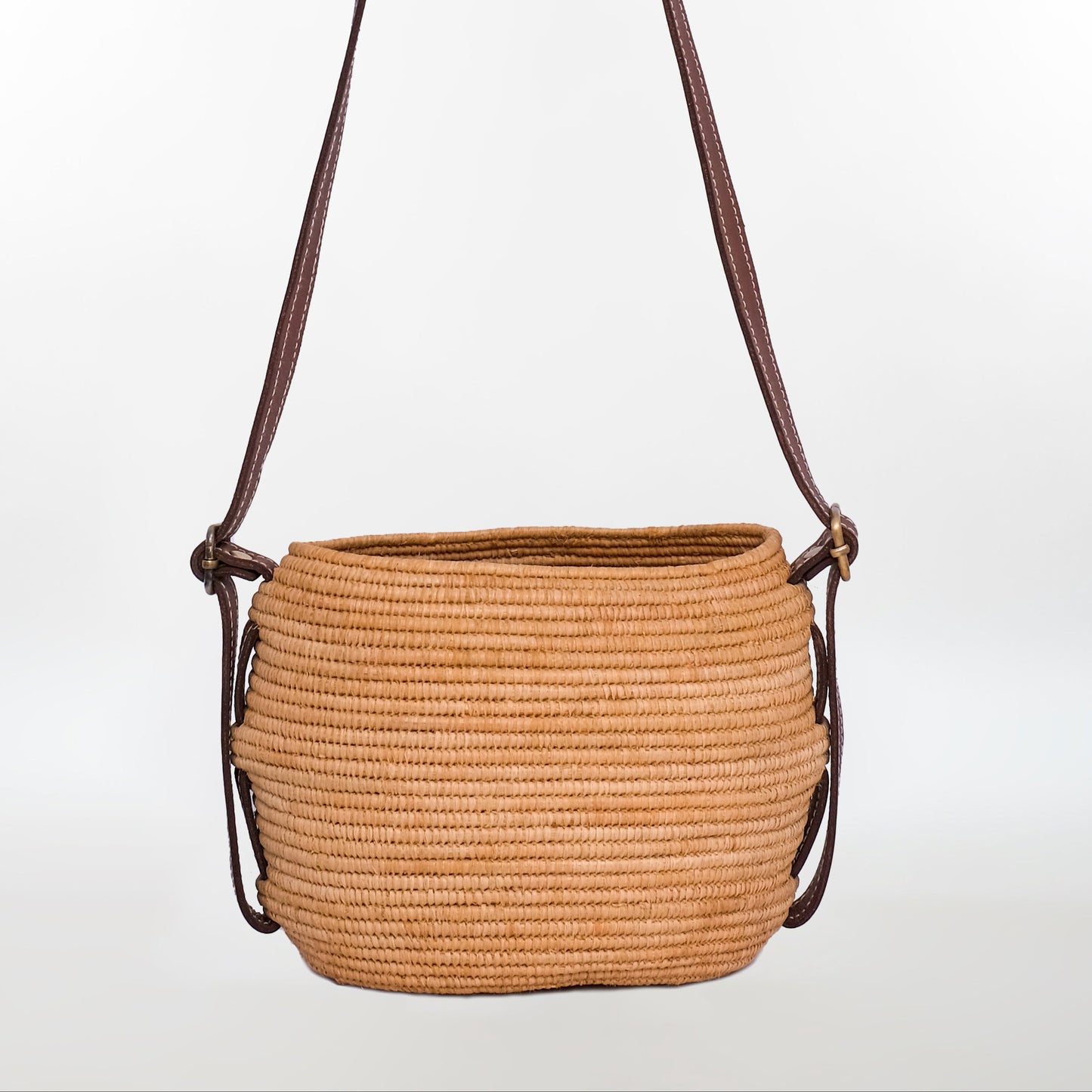 Natural raffia crossbody bag with dark brown leather shoulder strap and minimalist rounded shape