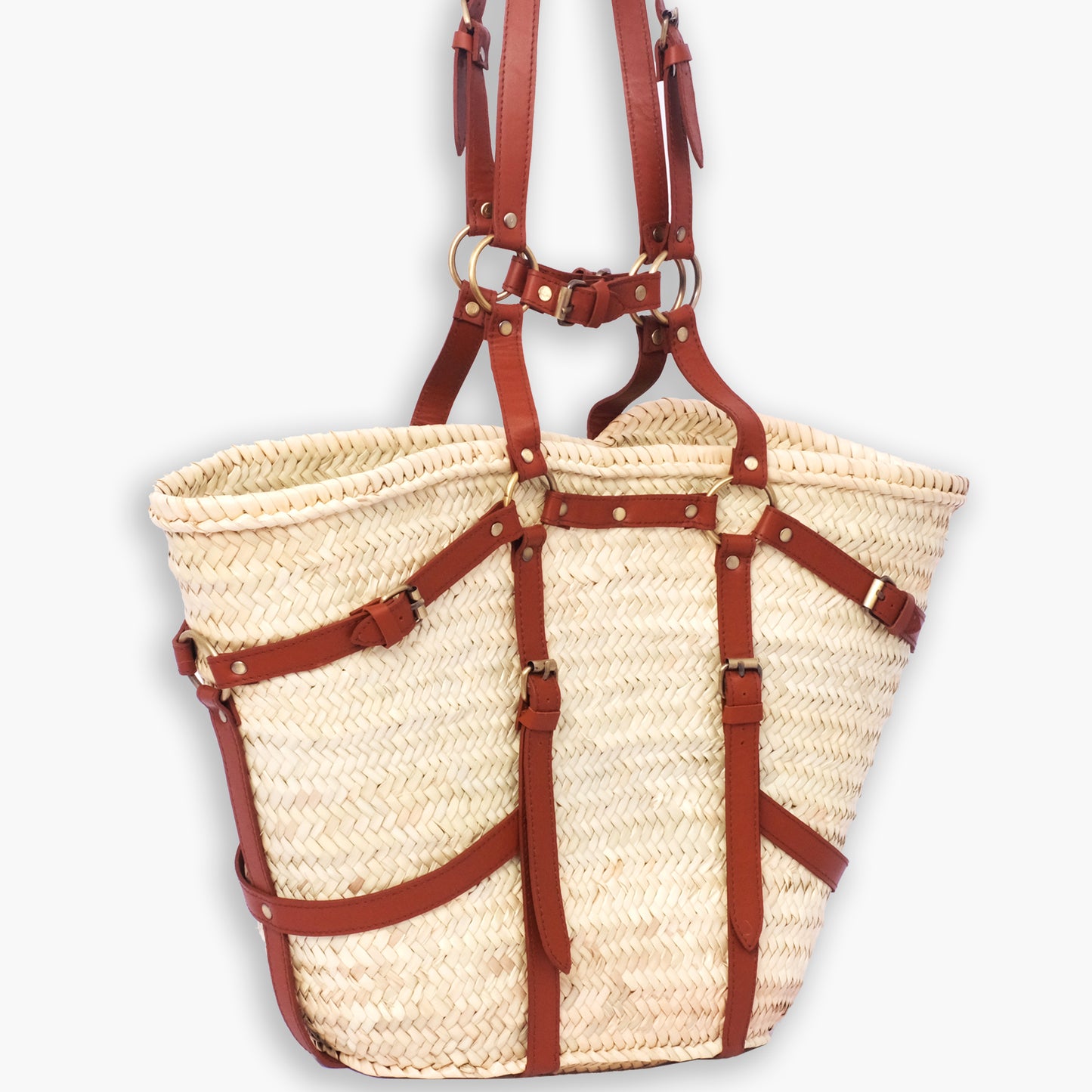 Moroccan straw tote with full harness-style leather straps, buckles, and brass hardware, photographed against a white background.