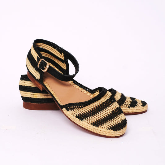 Women’s ankle-strap sandals handwoven from black and natural raffia, featuring striped pattern, closed heel, buckle closure, and leather sole, handmade in Morocco.